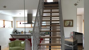 A modern interior with a central staircase, a dining area with red chairs, and a kitchen in the background.