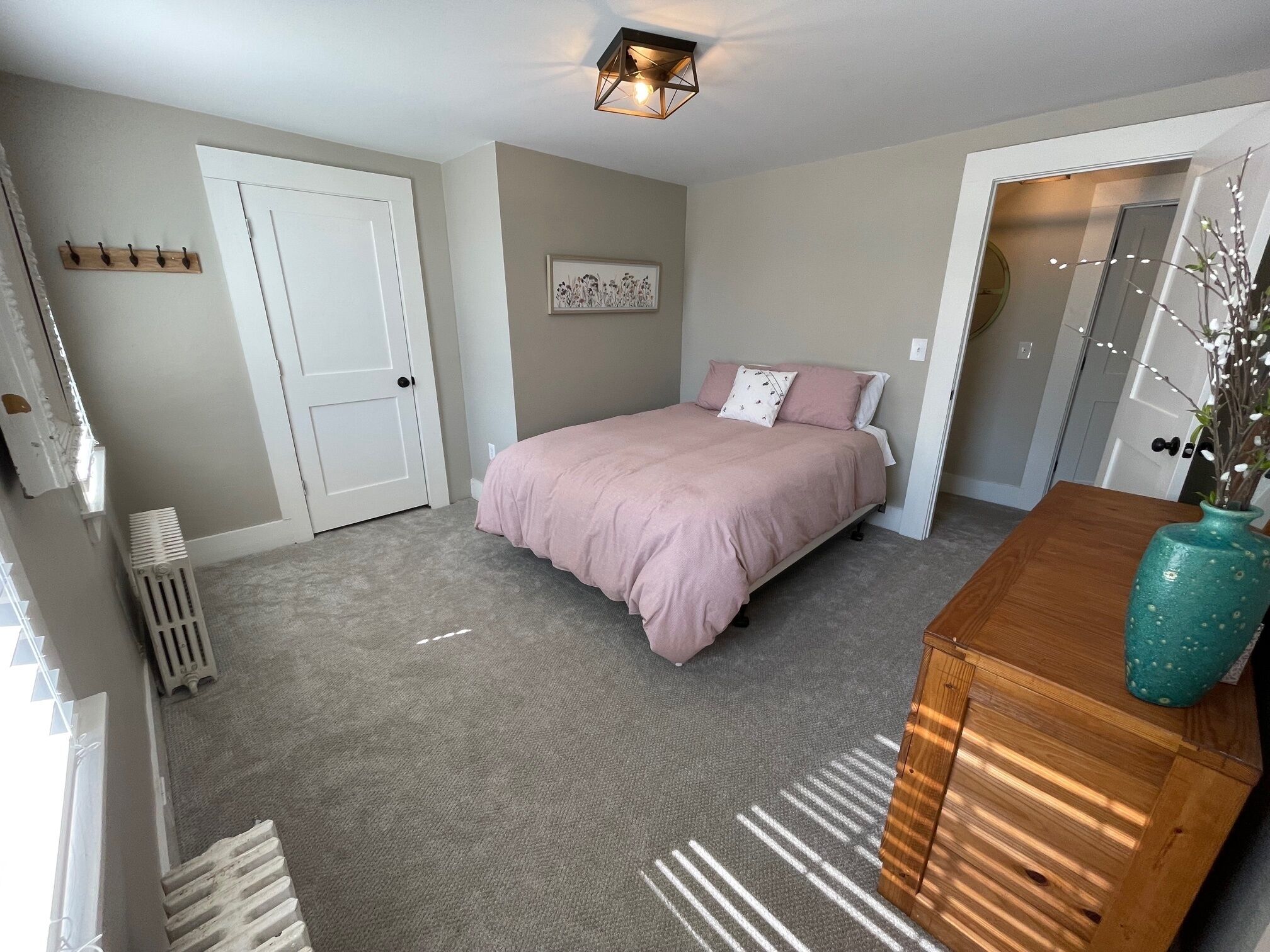 Un dormitorio con cama, cómoda de madera, radiador, puerta blanca y aplique de pared.