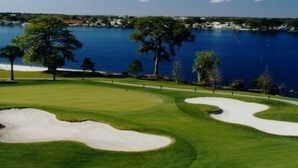 Ein Golfplatz mit Sandbunkern, einem Wasserhindernis und einer entfernten Küstenlinie mit Gebäuden.