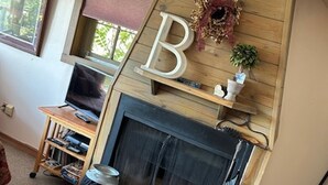 A living room with a fireplace, a wooden bookshelf, and a large letter 'B' decoration. - Mountain Top Eureka Springs Cabin with Full Glass Front Best Views in Eureka (Eureka Springs)