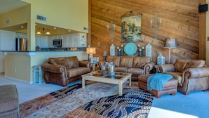 A spacious living room with leather sofas, a coffee table, and a patterned rug. A kitchen is visible in the background. - WATERFRONT CONDO  WITH AMAZING VIEW OF SUNRISE, SUNSET AND EXPANSIVE OPEN WATERS (Montgomery)
