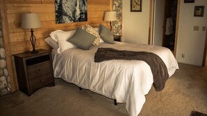 Quarto com cabeceira de madeira, uma cama grande com roupa de cama branca, duas luminárias de cabeceira e um quadro na parede.