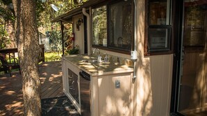 Área de cozinha externa pequena e coberta, com pia e janela, rodeada por um deck de madeira e uma árvore.