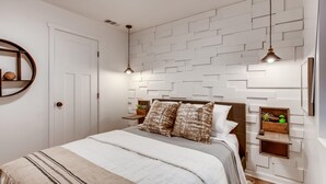 Un dormitorio con cama, mesitas de noche, estante en la pared y una puerta.