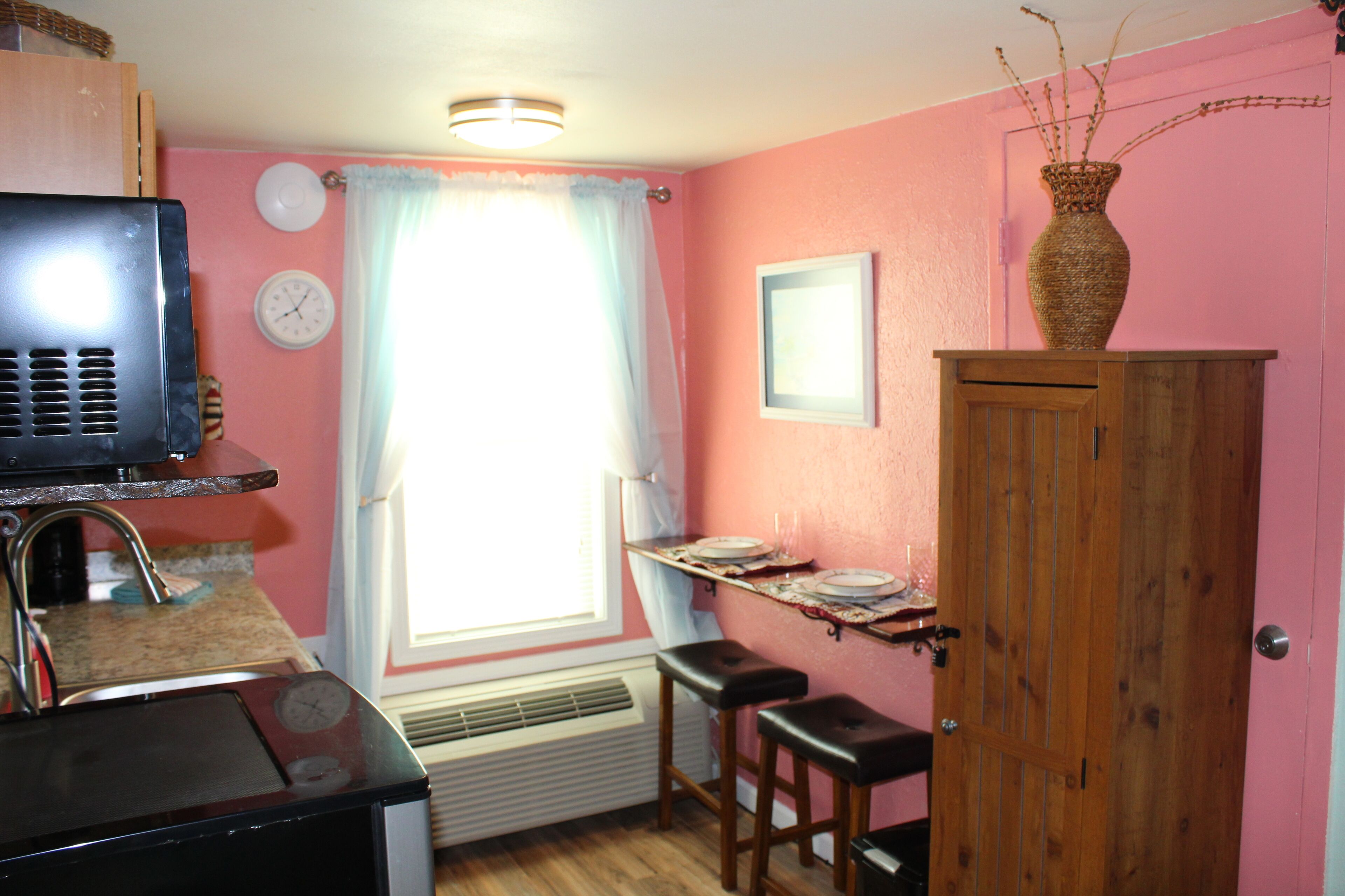 Une cuisine avec des murs roses, un meuble en bois, une fenêtre avec des rideaux, une horloge et un four à micro-ondes.