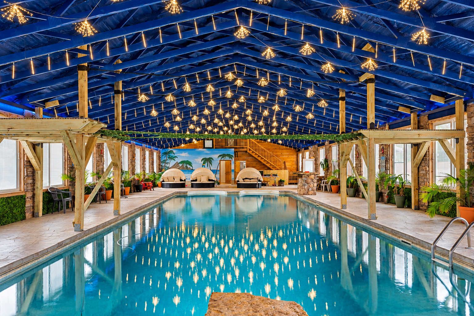 An indoor swimming pool with a ceiling adorned with hanging lights, wooden beams, and a tropical mural in the background.