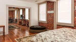 Une chambre avec un mur en briques, de grandes fenêtres, un lit recouvert d’une courtepointe à motifs floraux, un plancher en bois et un petit pouf.
