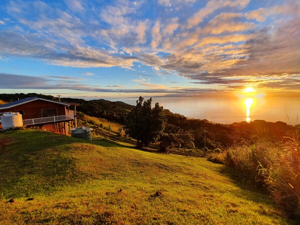 Ein Haus mit Balkon mit Blick auf einen grasbewachsenen Hügel und einen Sonnenuntergang über dem Wasser.