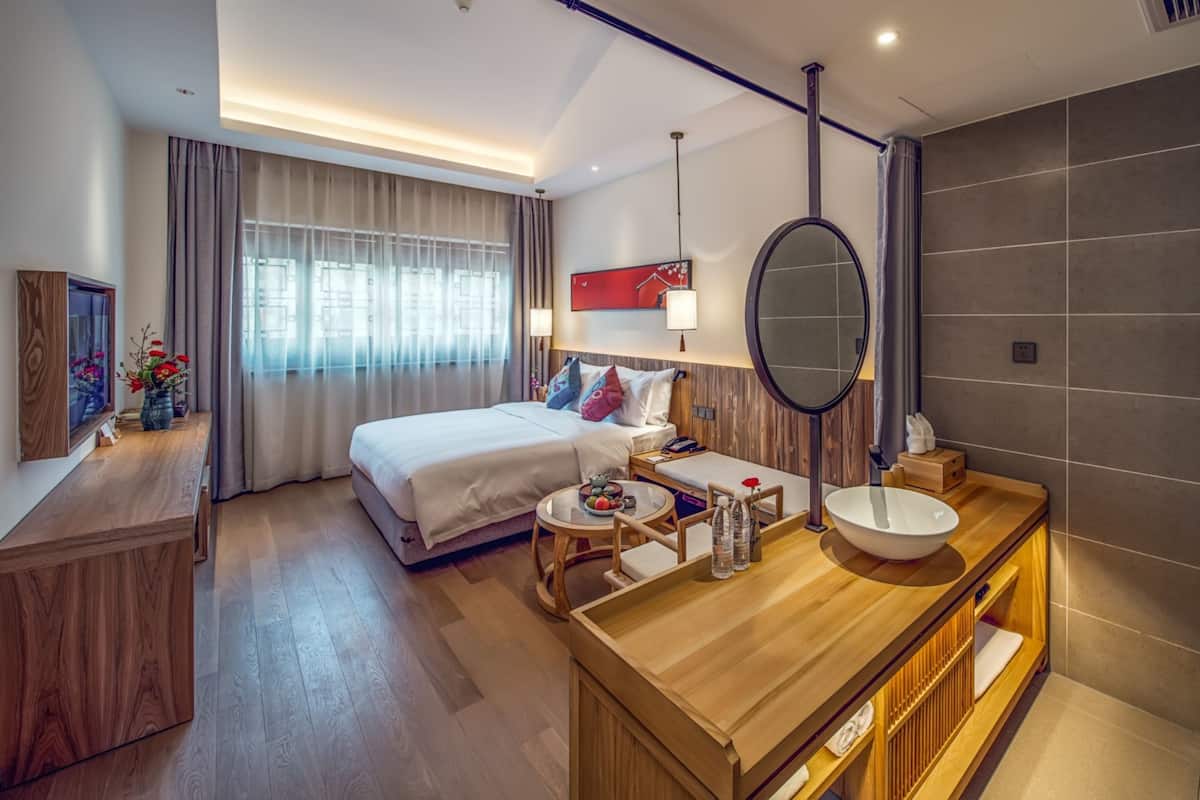 A modern hotel room with a large bed, a wooden vanity with a mirror, and a large window with curtains.