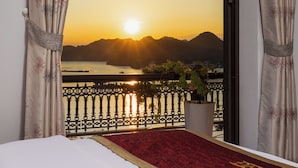 A balcony with a view of the sunset over water and hills, a red carpet, and a potted plant.
