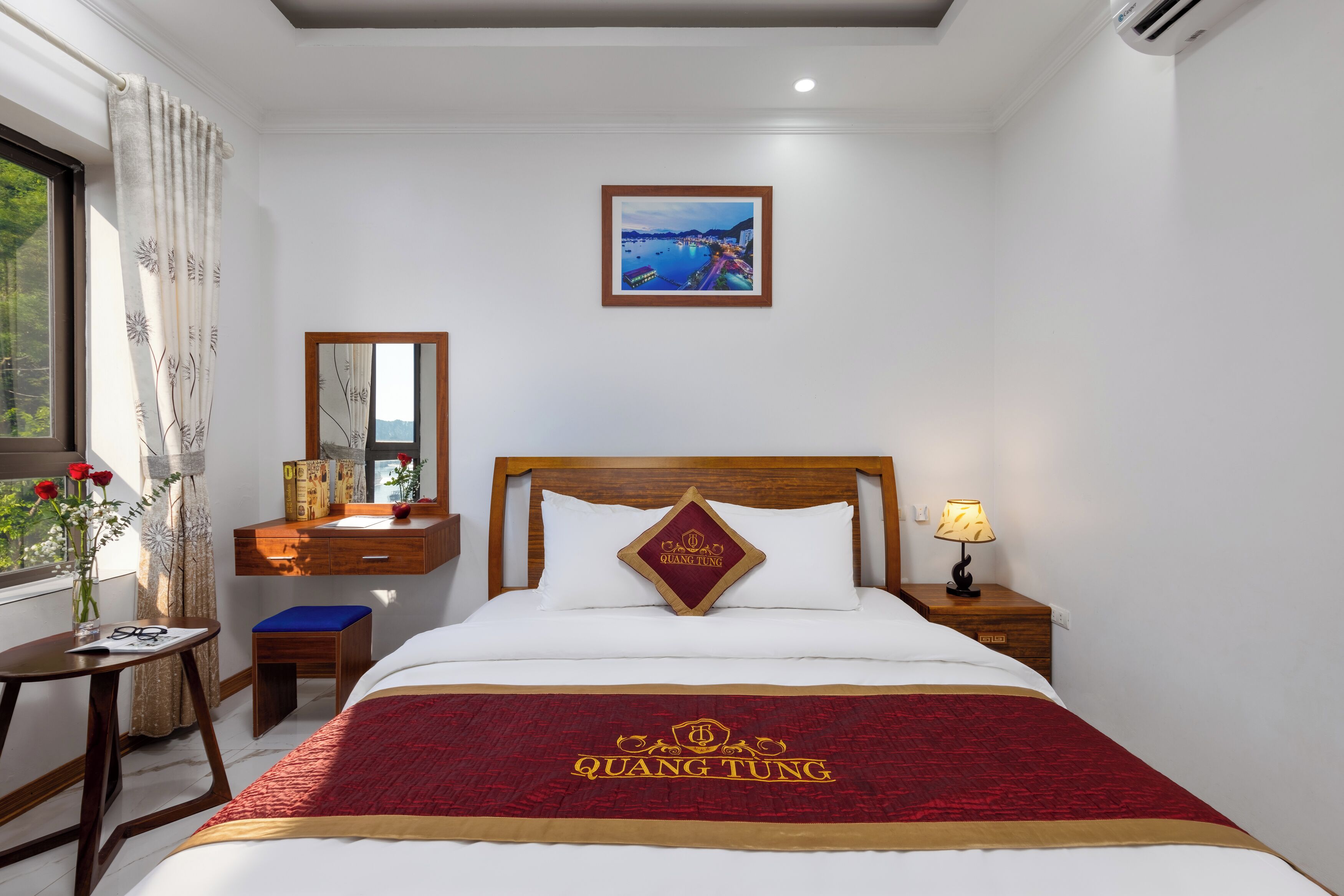 A hotel room with a bed, a nightstand with a lamp, a desk with a chair, a window with curtains, and a framed picture on the wall.