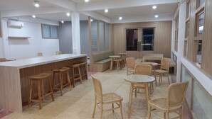 A modern cafe with a long counter, bar stools, round tables, and wicker chairs. - Sataya Apartment (Maha Sarakham)