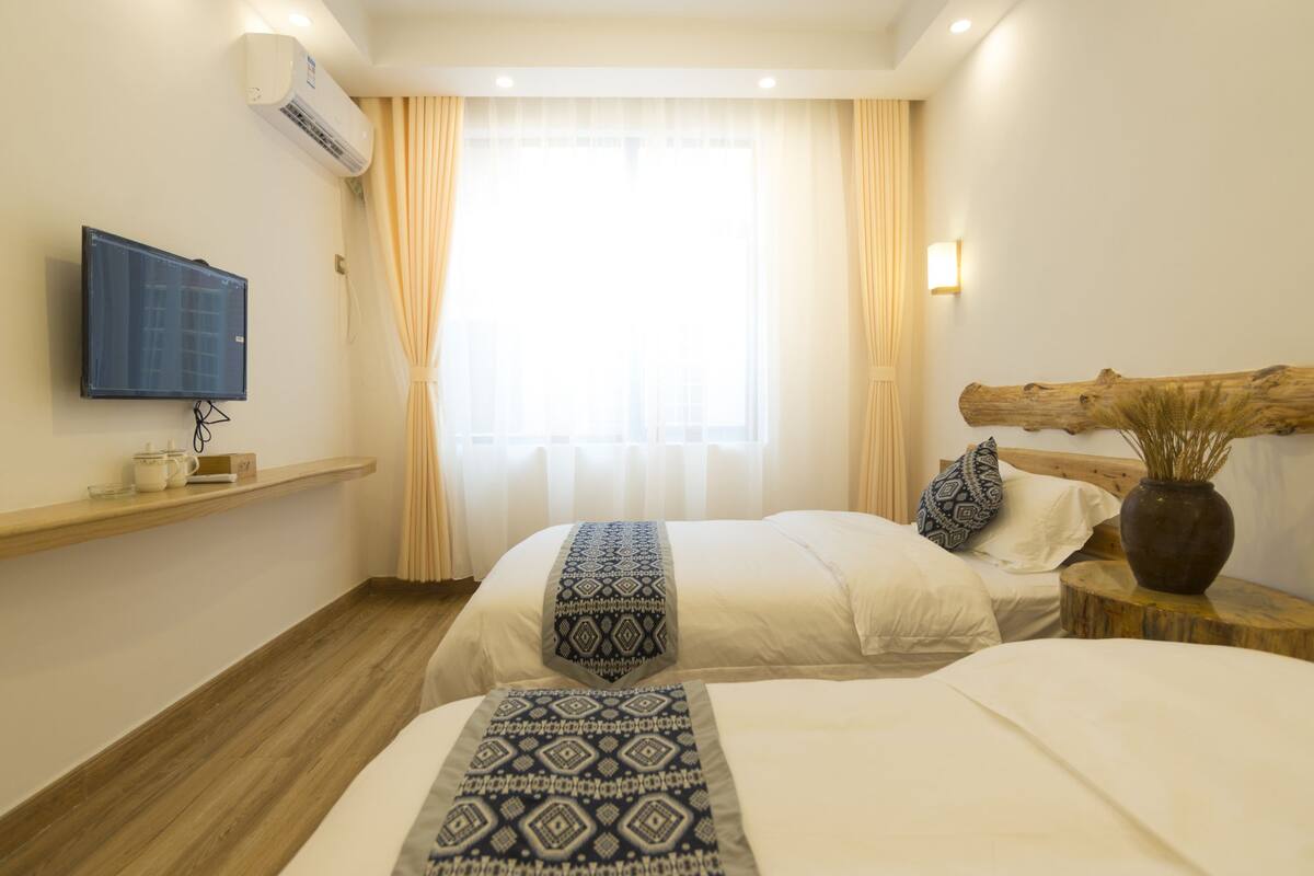 Una habitación de hotel con dos camas, un televisor montado, un cabecero de madera, un florero con plantas secas y una ventana con cortinas translúcidas.
