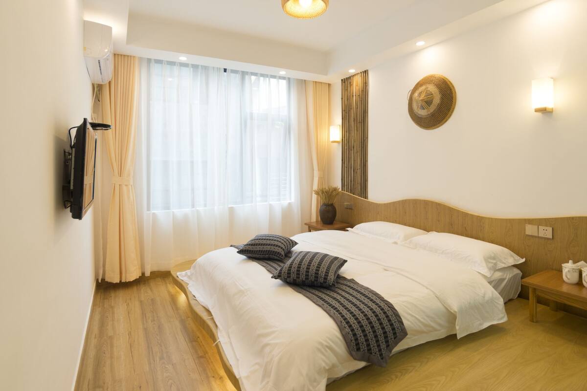 Una habitación de hotel con una cama grande, una mesita de noche, un televisor montado en la pared y una ventana con cortinas traslúcidas.