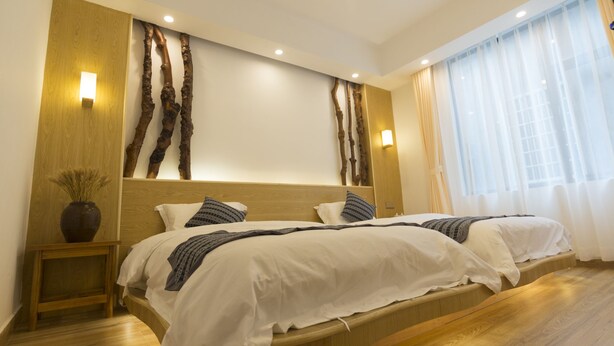 A modern bedroom with a wooden headboard, two beds, bedside tables, and a large window with sheer curtains.