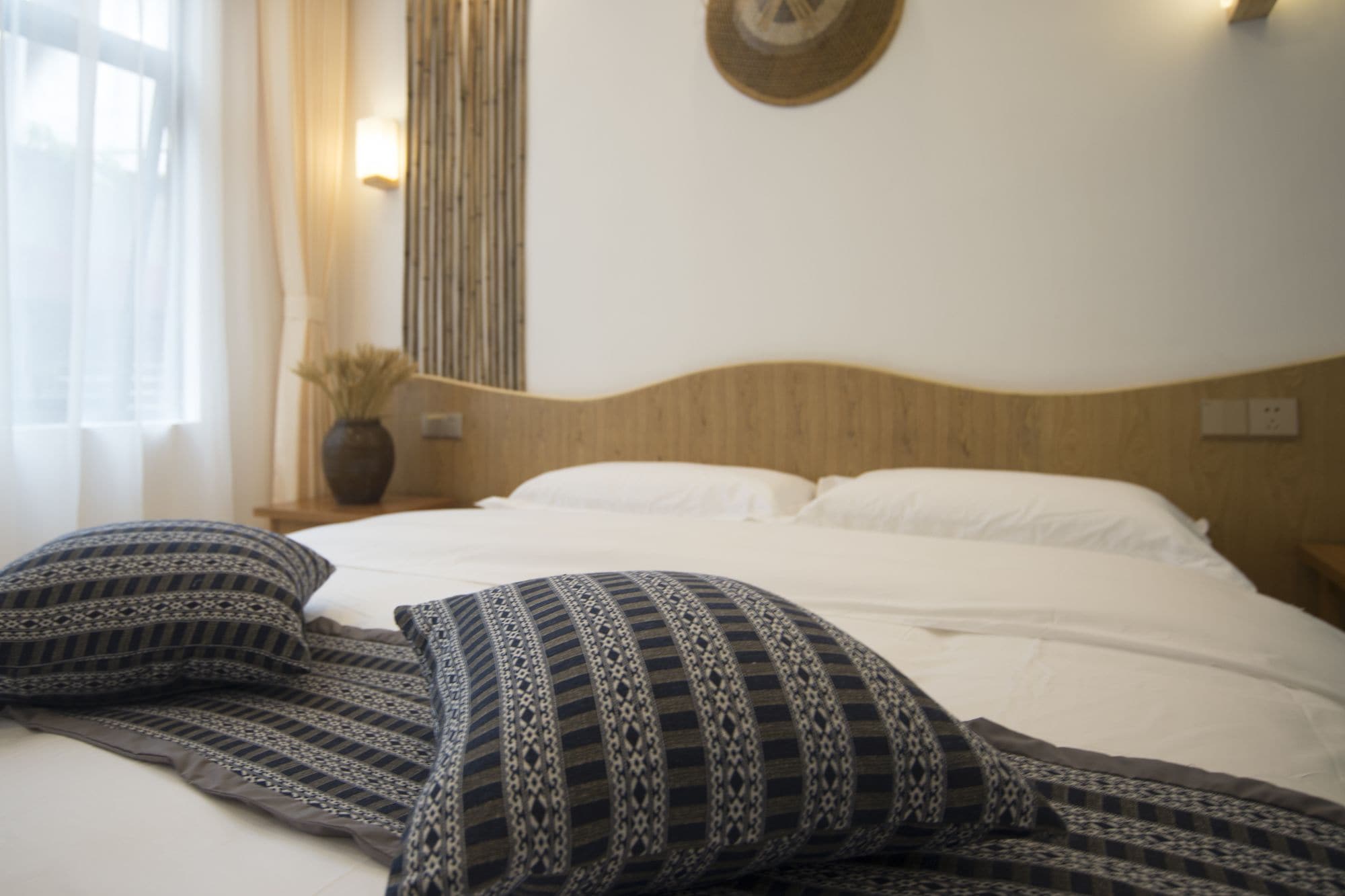 A neatly made bed with patterned pillows, a wooden headboard, a wall-mounted light fixture, and a vase with dried plants on a bedside table.