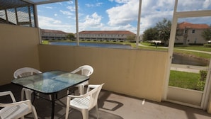 Un patio cubierto con una mesa de vidrio y sillas blancas, con vista a un patio interior con edificios y un cuerpo de agua.