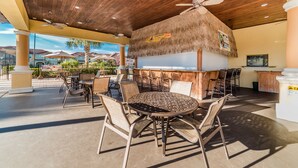 An outdoor patio with round tables and chairs, a thatched roof, and a pool area in the background. - Ip60387 - Paradise Palms - 5 Bed 4 Baths Townhome (Kissimmee)