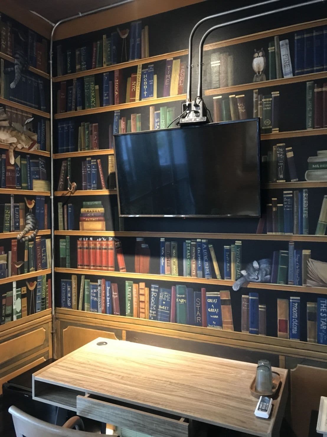A library with bookshelves, a flat-screen TV mounted on the wall, and a wooden table with a remote control.