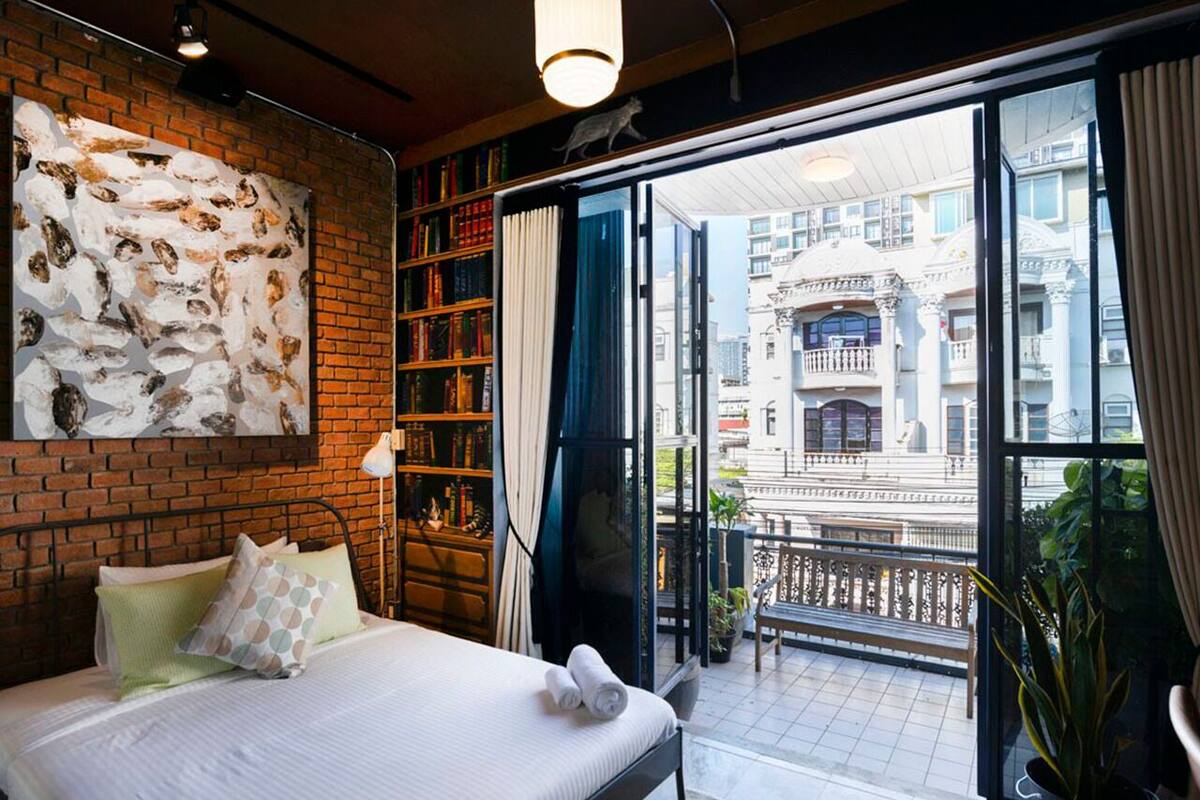 Une chambre d’hôtel avec un lit, une bibliothèque, une grande fenêtre donnant sur des immeubles et un balcon.