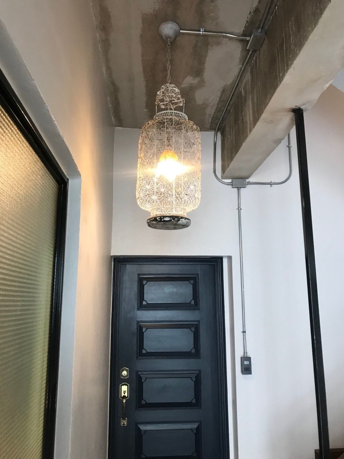 A black door with a keyhole and a lock, a hanging light fixture, and a ceiling with exposed pipes.