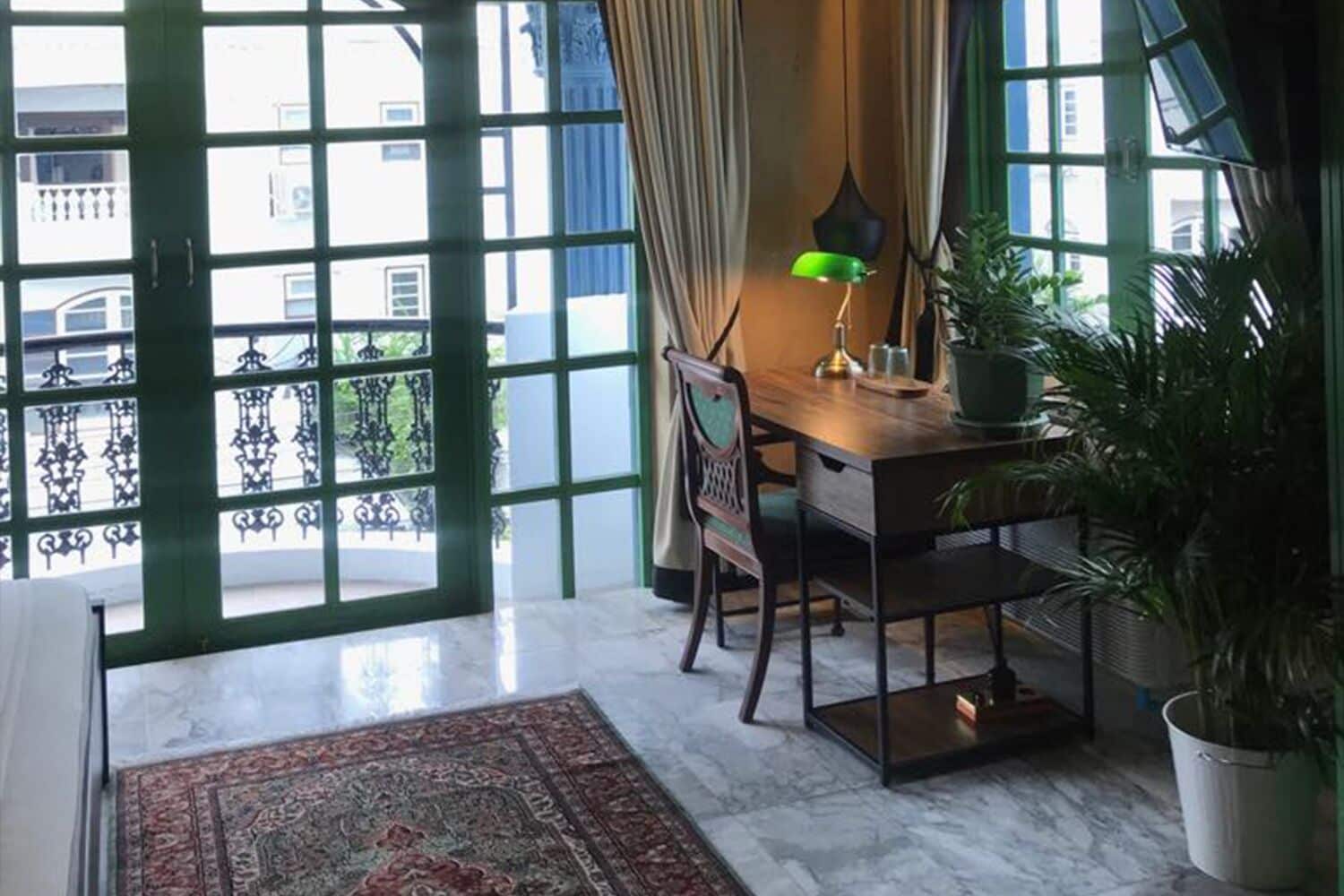 A room with a large window, a desk with a lamp, a chair, a rug, and a potted plant.