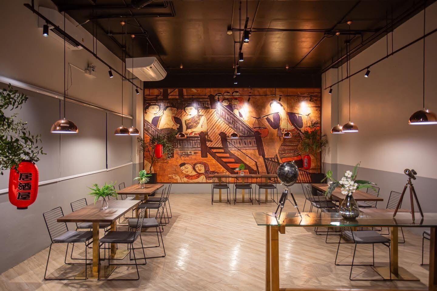 Un espace repas moderne avec des tables et des chaises en bois, une fresque murale au fond et des luminaires suspendus.