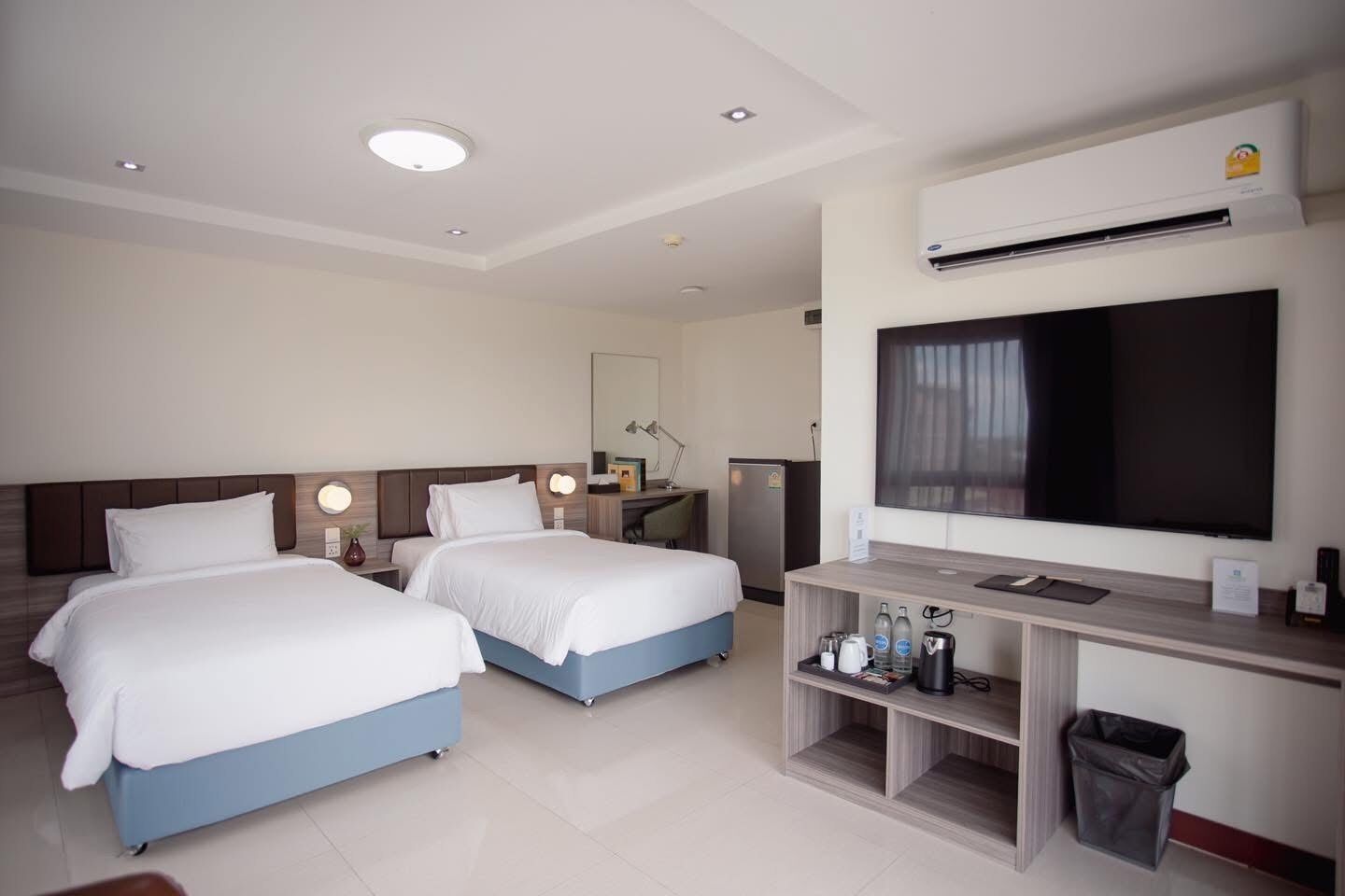 A hotel room with two beds, a desk with a flat-screen TV, a minibar, and a trash bin.