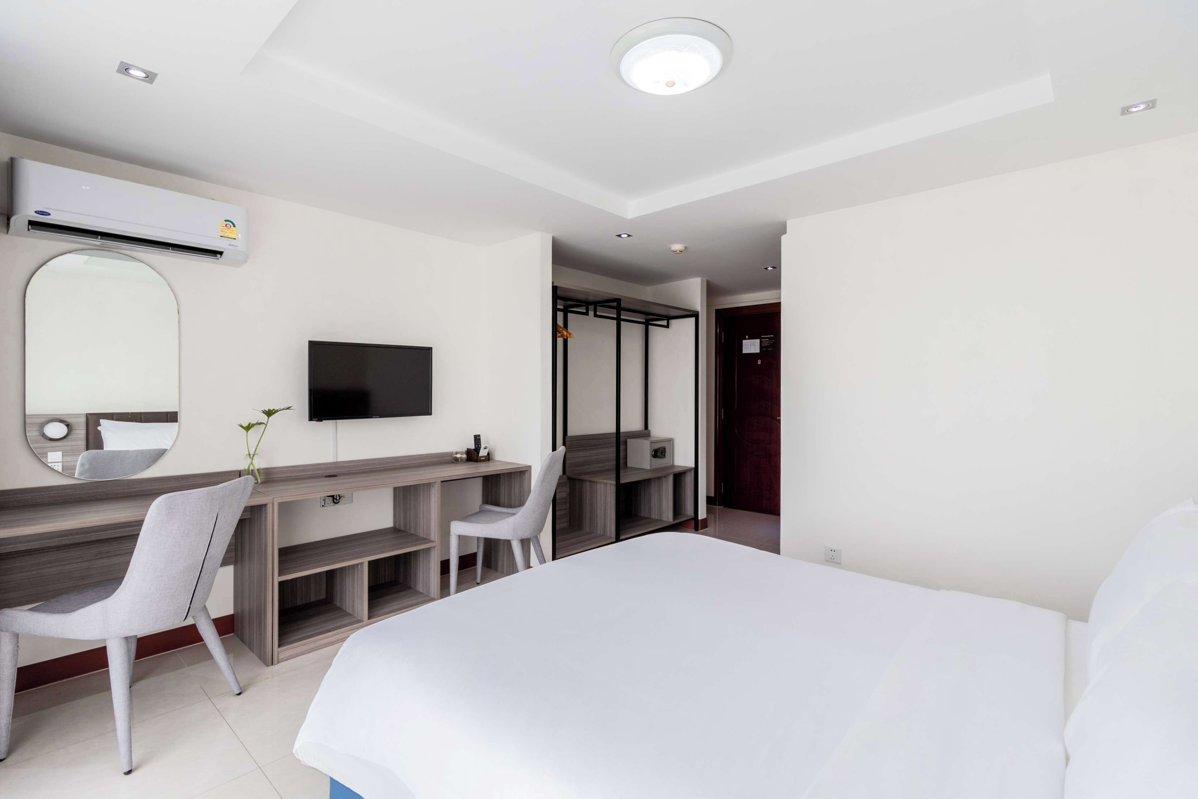 A modern hotel room with a bed, a desk with a chair, a TV, and an air conditioning unit.