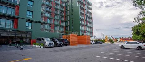 Un hotel de varios pisos con fachada verde, estacionamiento y una estatua al fondo.