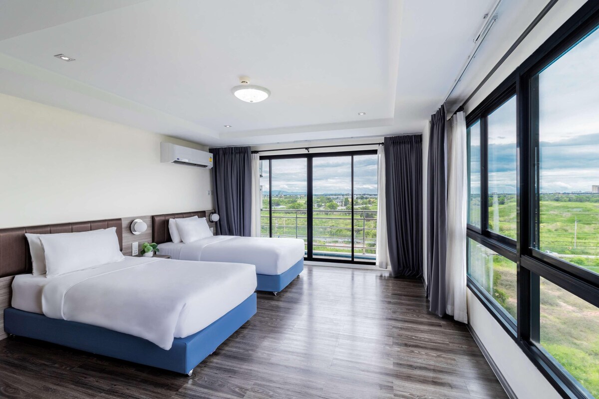 A hotel room with two beds, a large window with a view, a ceiling light, and an air conditioning unit.