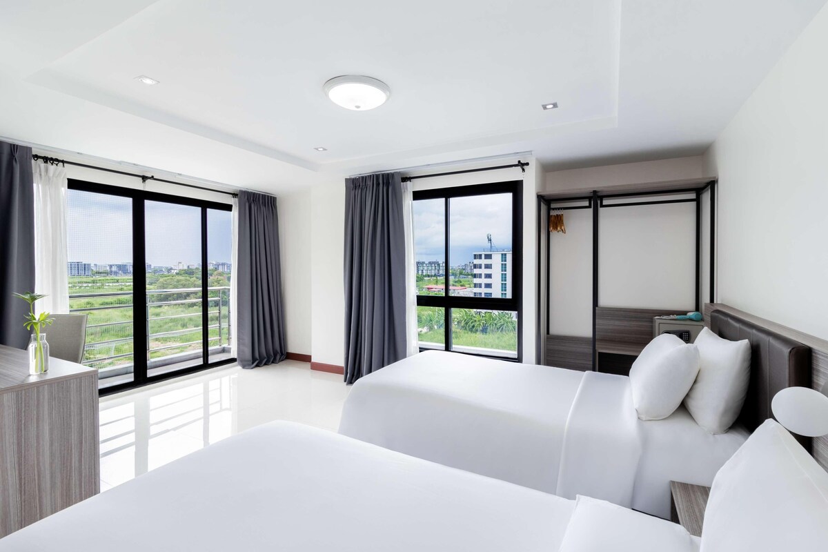 A hotel room with two beds, large windows with curtains, a desk with a vase, and a view of buildings.