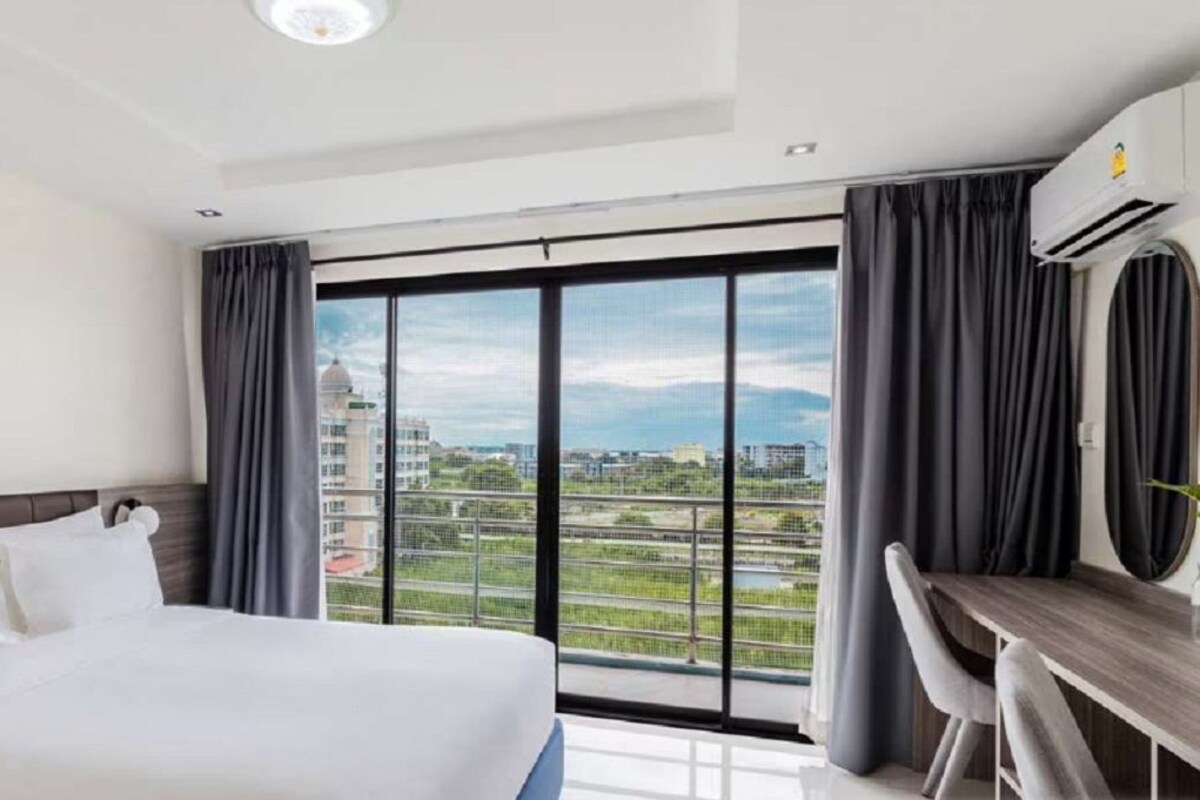 A hotel room with a large bed, a desk with a chair, and a view of a cityscape through large windows.