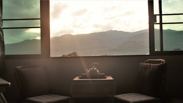 A room with a view of mountains through a window, a small table with a teapot, and two cushioned chairs. - 5 Min Ninman Road & Best Mountain View (Chiang Mai)