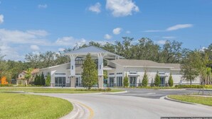 A modern building with a covered entrance and large windows, surrounded by greenery and a curved road. - Aco240198 - Golden Palms Resort - 7 Bed 6 Baths Villa (Kissimmee)