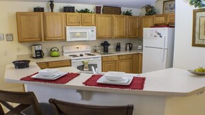 Una cocina con armarios de madera, un microondas blanco, una estufa y un refrigerador. Hay una zona de comedor con una mesa y sillas.