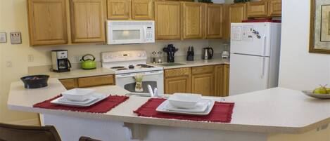 A kitchen with wooden cabinets, a white microwave, a stove, and a refrigerator. There is a dining area with a table and chairs.