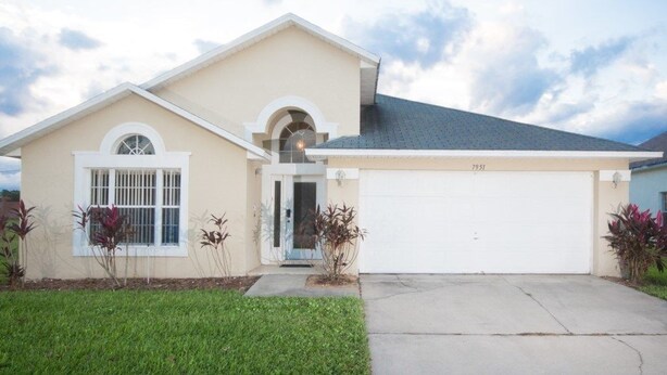 A single-story house with a beige exterior, a white garage door, and a small front yard with plants. - Ip60224 - Rolling Hills Estates - 4 Bed 3 Baths Villa (Kissimmee)