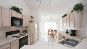 Una cocina moderna con armarios de madera clara, un microondas negro, una estufa y un fregadero. Hay una zona de comedor con una mesa y sillas, y plantas sobre los armarios.