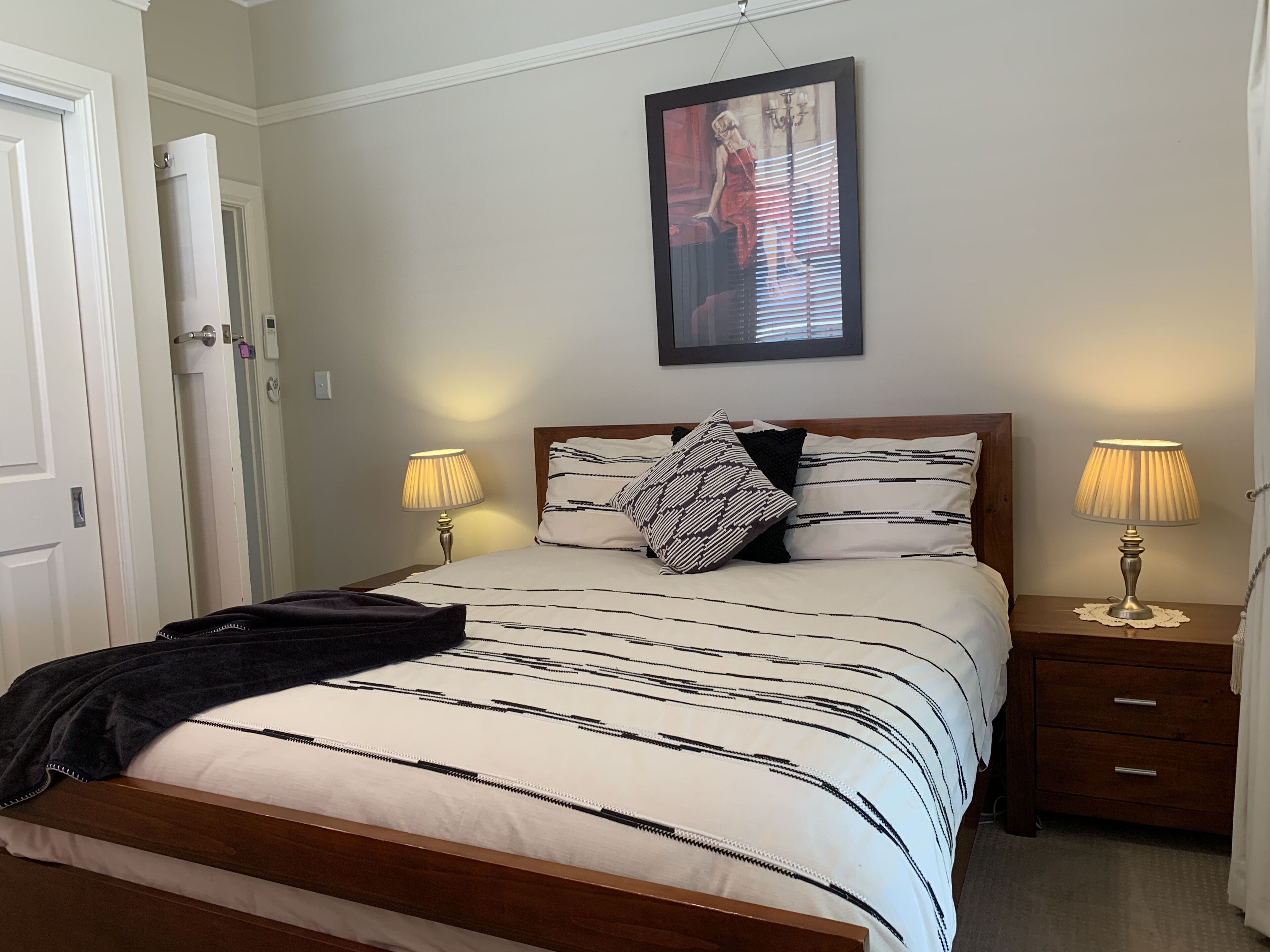 Een net opgemaakt bed met strepen op het beddengoed, twee bedlampjes, een ingelijste foto aan de muur en een houten nachtkastje met lades.