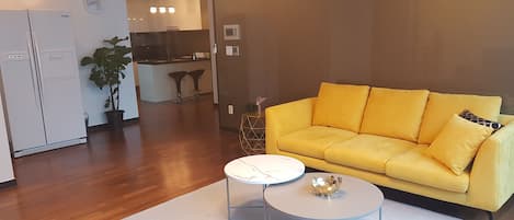 Un salon moderne avec un canapé jaune, une table basse blanche et une cuisine en arrière-plan.