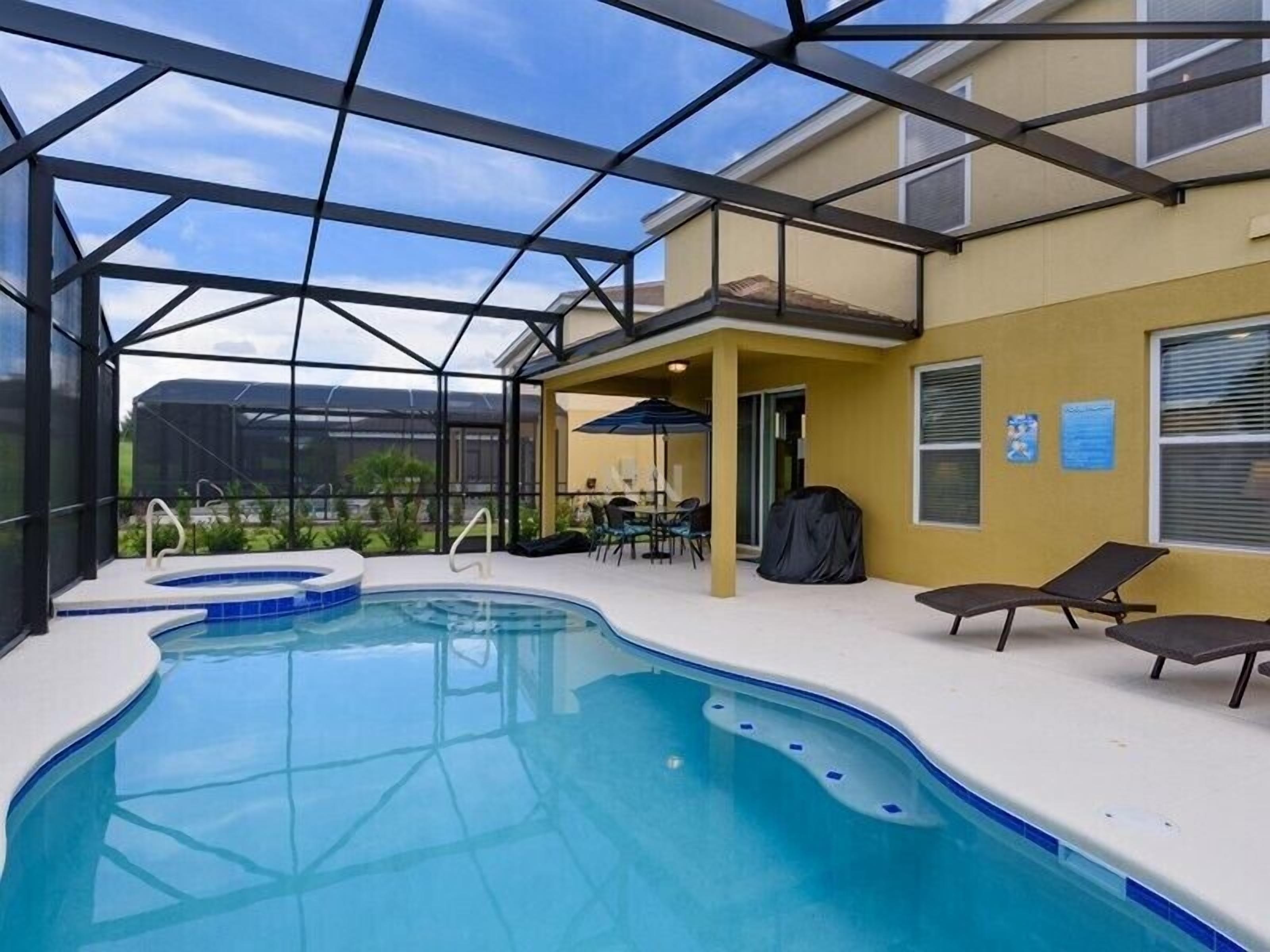 A covered swimming pool with a hot tub, lounge chairs, and a patio area with a table and chairs.