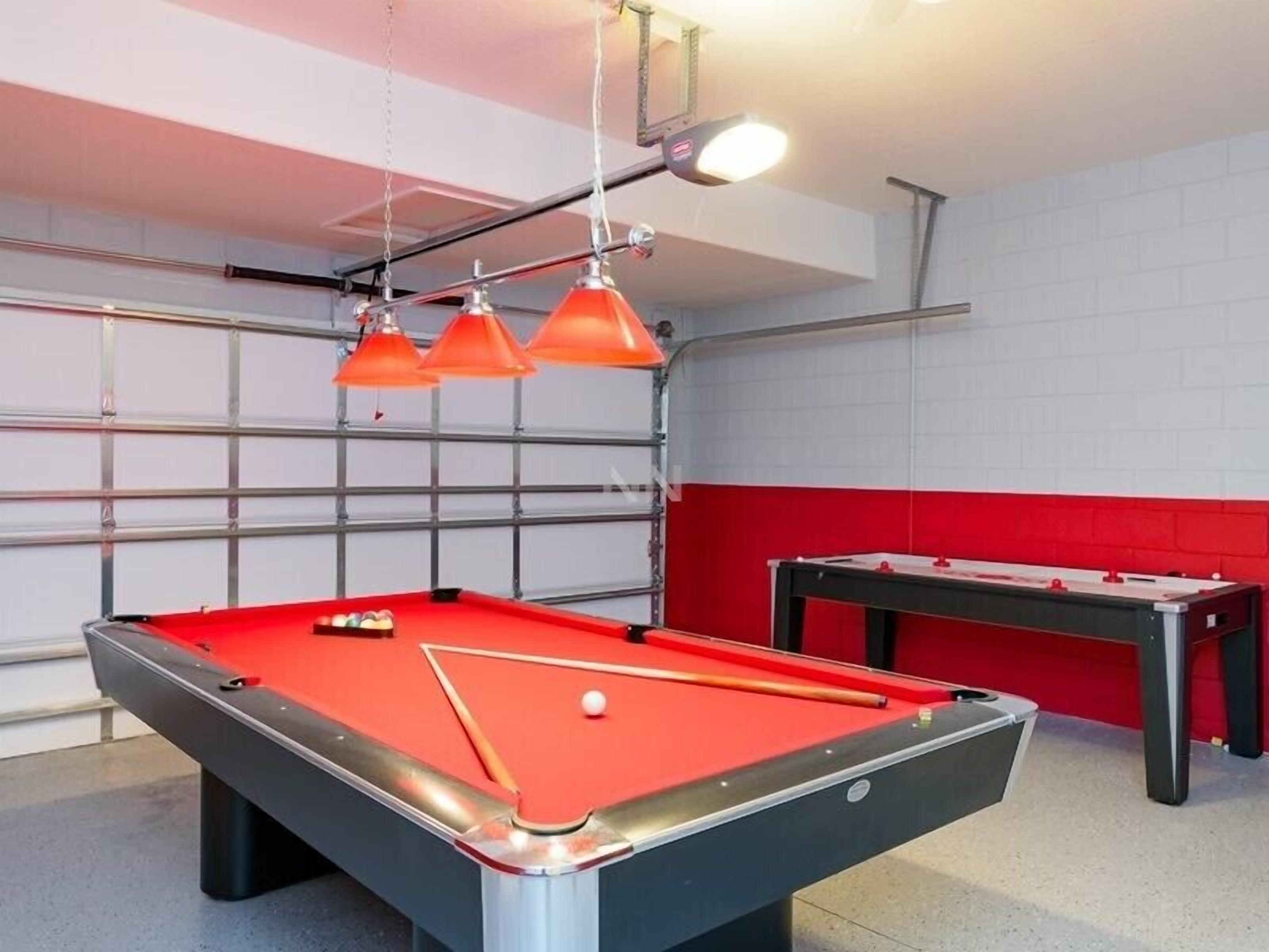 A pool table with three orange lamps above it, a red and black pool table, and a red padded wall in a room with a white ceiling and metal grid.