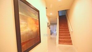 A hallway with a wooden staircase, a framed abstract painting on the wall, and a glimpse of a room with a window and curtains.