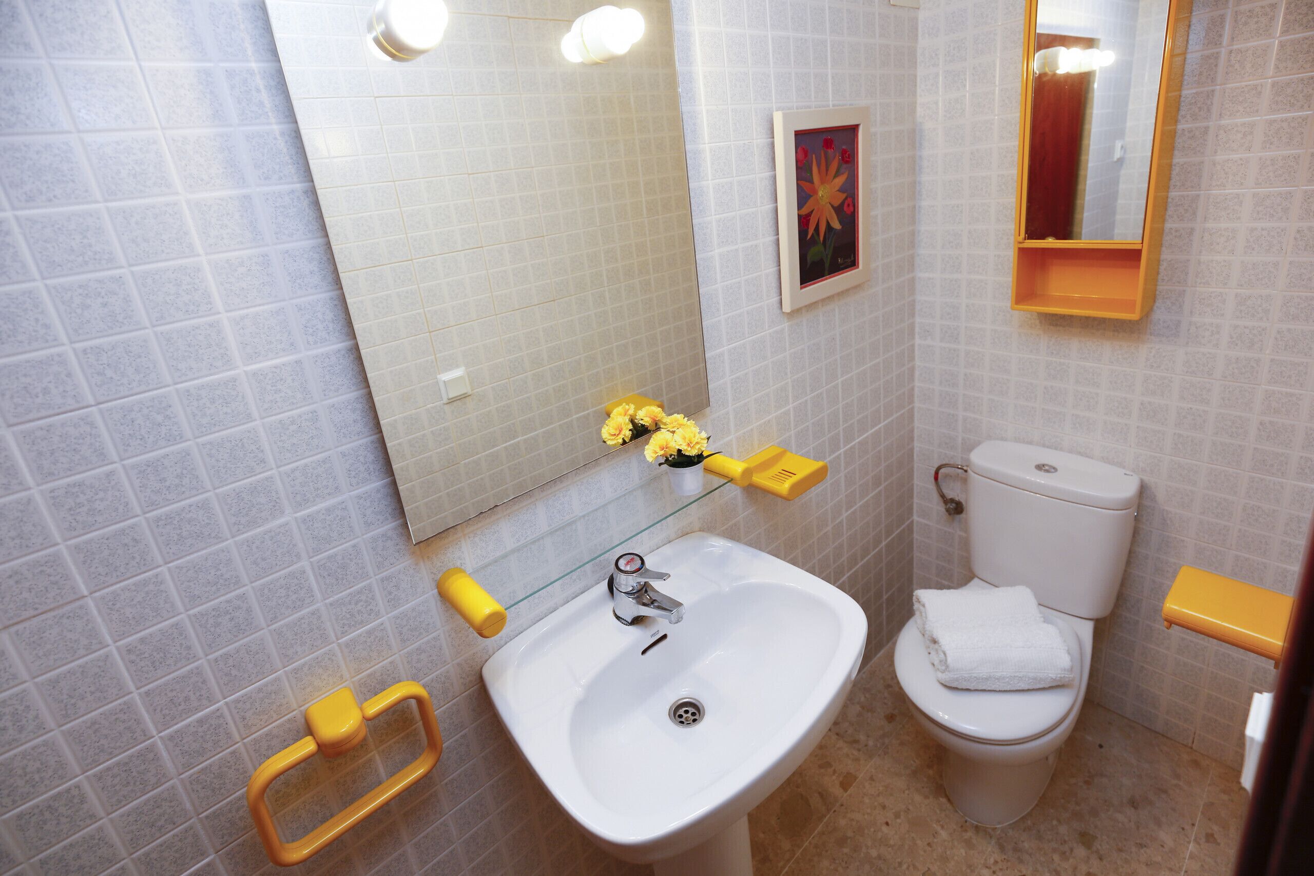 A bathroom with a white sink, a toilet, and a mirror. There are yellow soap dispensers and a framed picture on the wall.