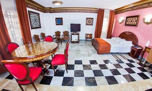 A hotel room with a checkered floor, a round dining table with chairs, a bed with a headboard, and a television. - Dar Diaf Cheraga (Algiers)