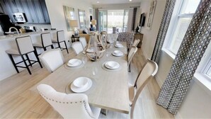 A dining area with a long table set for six, surrounded by white upholstered chairs. A kitchen with modern appliances is visible in the background. - Aco233851 - The Encore Club Resort - 6 Bed 6.5 Baths Villa (Kissimmee)