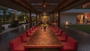 Een lange eettafel met rode, beklede banken, omringd door een overdekt terras met houten vloer en potplanten.