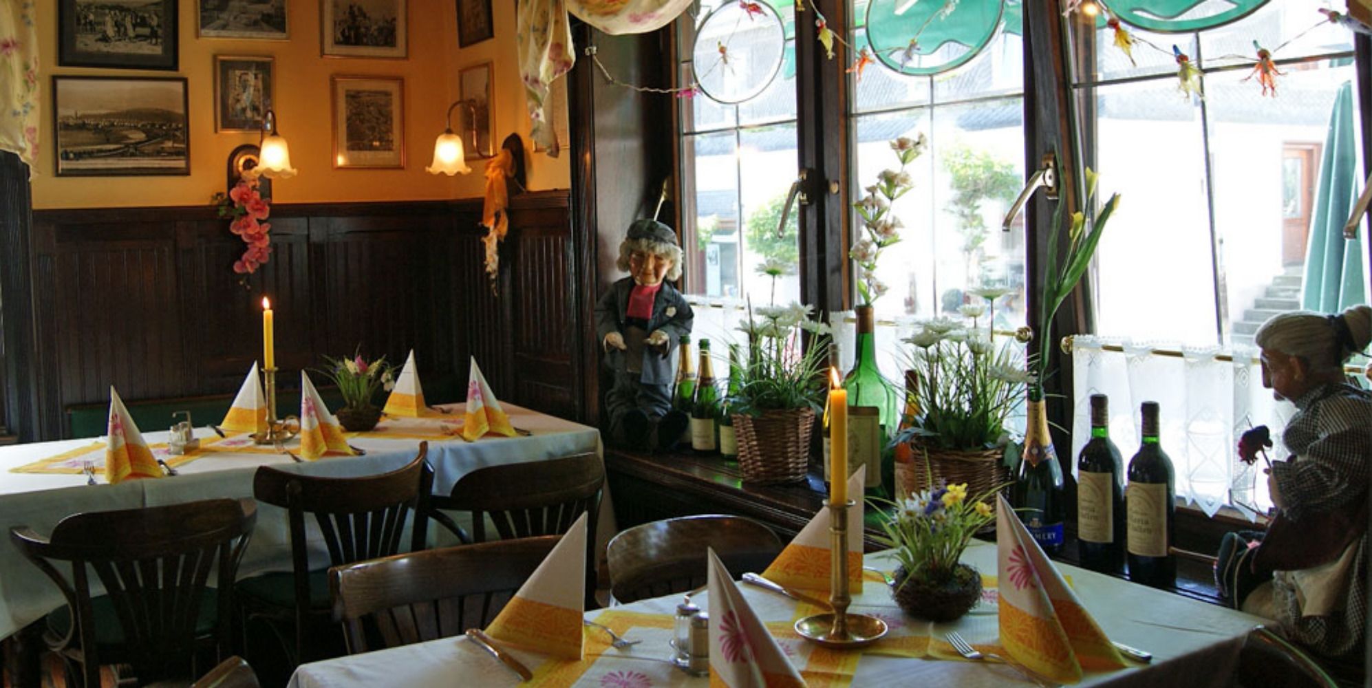 A restaurant with tables set for dining, featuring a traditional decor with framed pictures on the walls and a variety of plants and flowers on the tables.