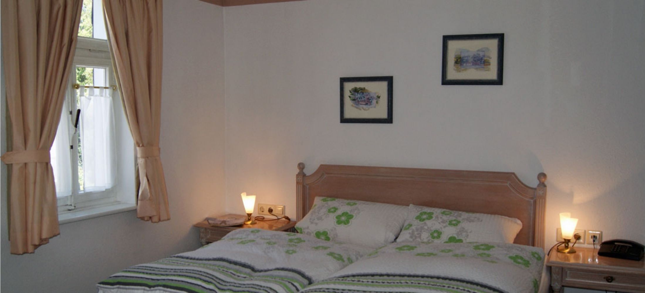 A bedroom with a single bed, two bedside tables, a window with curtains, and two framed pictures on the wall.